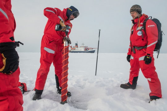 three scientists dressed in snow suits drill ice cores out on the frozen, white landscape, ship can be seen far away.