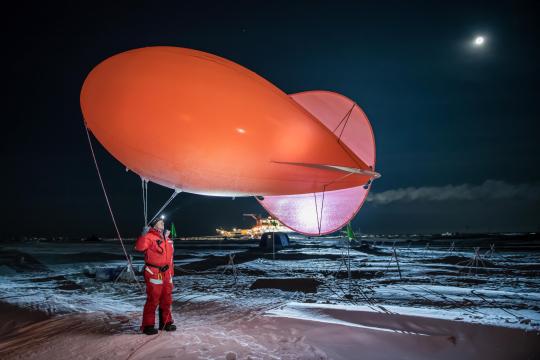 Known as Miss Piggy, the weather balloon collects data within the few thousand feet of atmosphere above the ice. Graeser wears a red snowsuit, and the ship Polarstern is visible in the distance. December 12, 2019. Photo: Esther Horvath