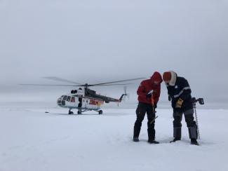 Jakob Belter and Jan Rohde of AWI determine the thickness of the ice floe equipped with drill and gauge. Photo: Katharina Weiss-Tuider / AWI Two scientists on ice, helicopter in background